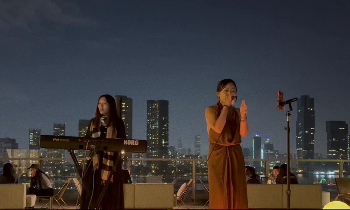 Jaya Lee singing on a rooftop terrace at night, the city lit behind her